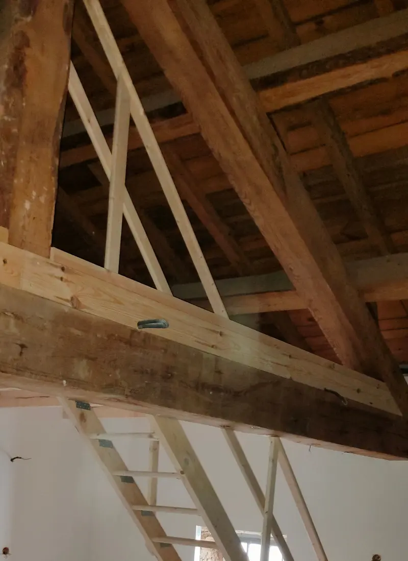 Charpente bois ancienne restaurée au-dessus d'une pièce aux murs blancs, avec mezzanine